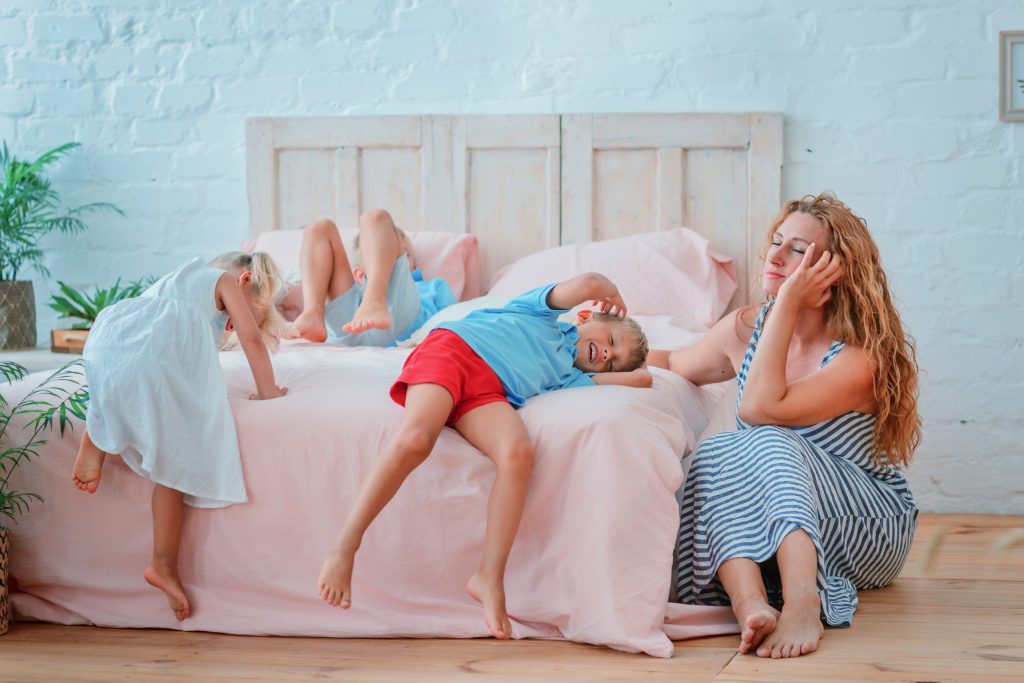 Mother with long fair hair, wearing a light blue cotton summer dress sits exhausted at the side of the bed where her two flopped out tired children are sprawled out, also wearing light cotton tops and shorts