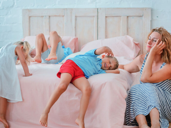 Sleepy mum with her three children after a day out, where the kids are sprawled out on the king-sized bed, they're all in their day clothes, the mum is slumped exhausted at the side of the bed.