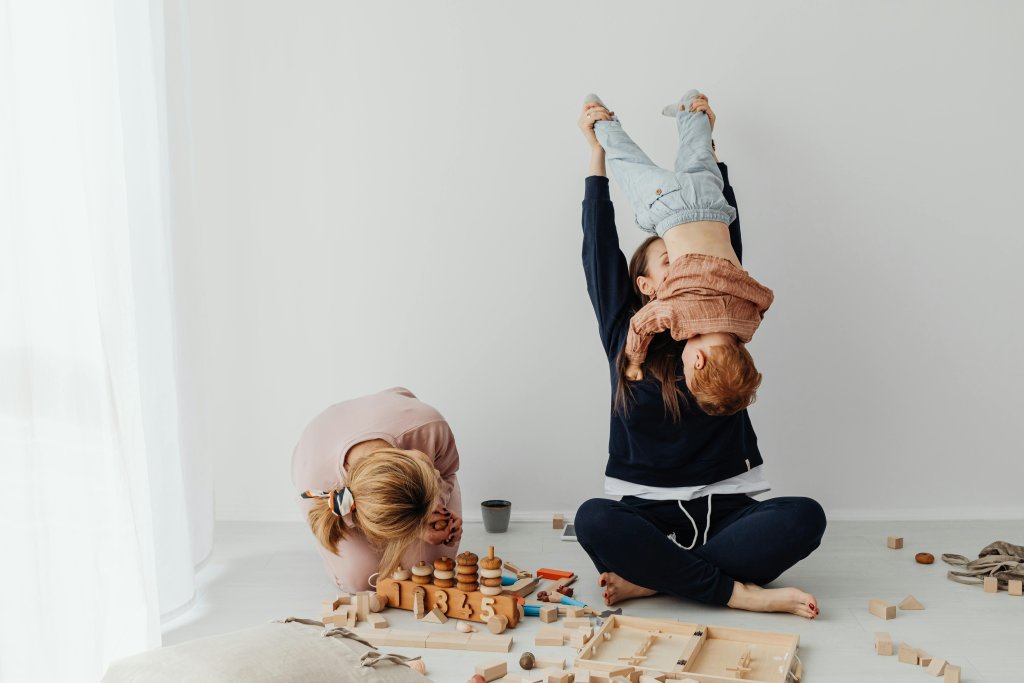 Mum on the floor playfully holding a toddler upside down with another child playing on the floorl