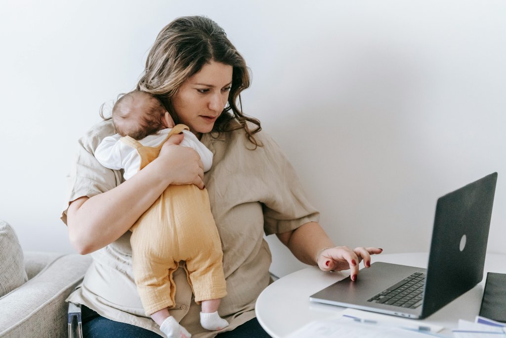 Mum with baby slumped over her shoulder while trying to use the laptop with the free hand.
